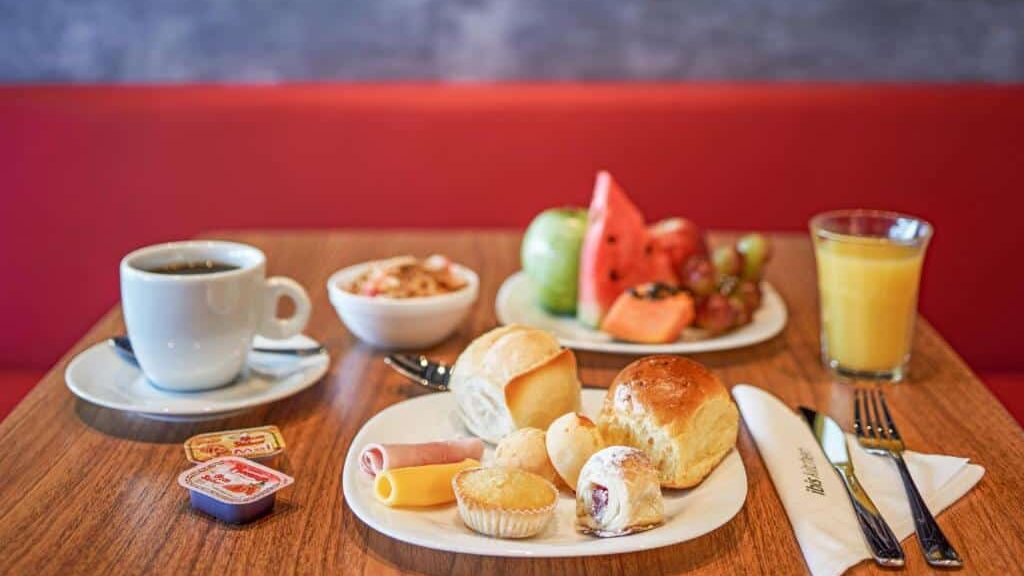 Mesa de pequeno-almoço do Hotel Ibis Lisboa José Malhoa com café, cereais, fruta, pão, queijo, fiambre, compota e sumo de laranja.