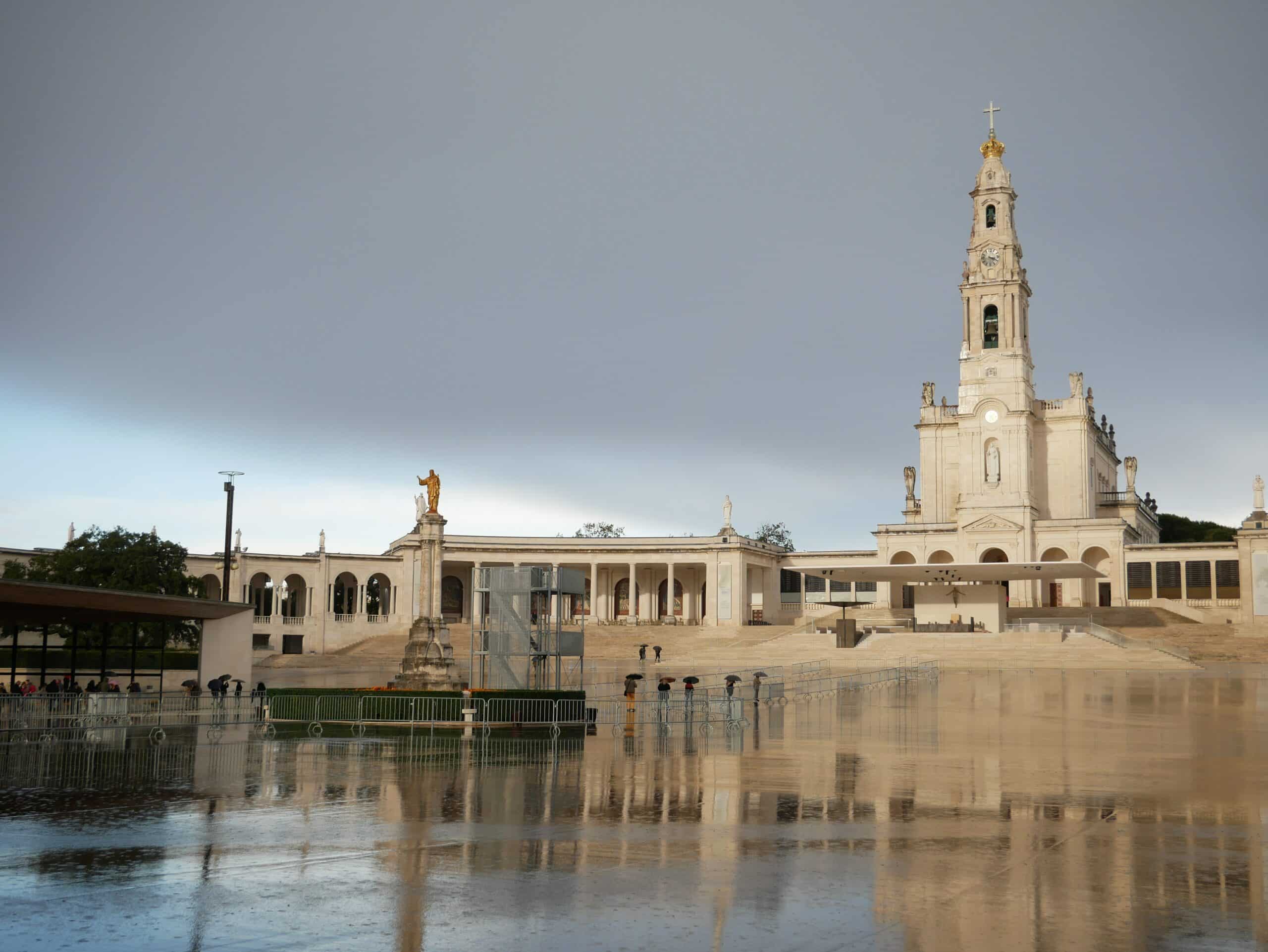 Grande basílica com uma alta torre sineira, vista de uma praça molhada e refletora sob um céu nublado, no Santuário de Fátima.