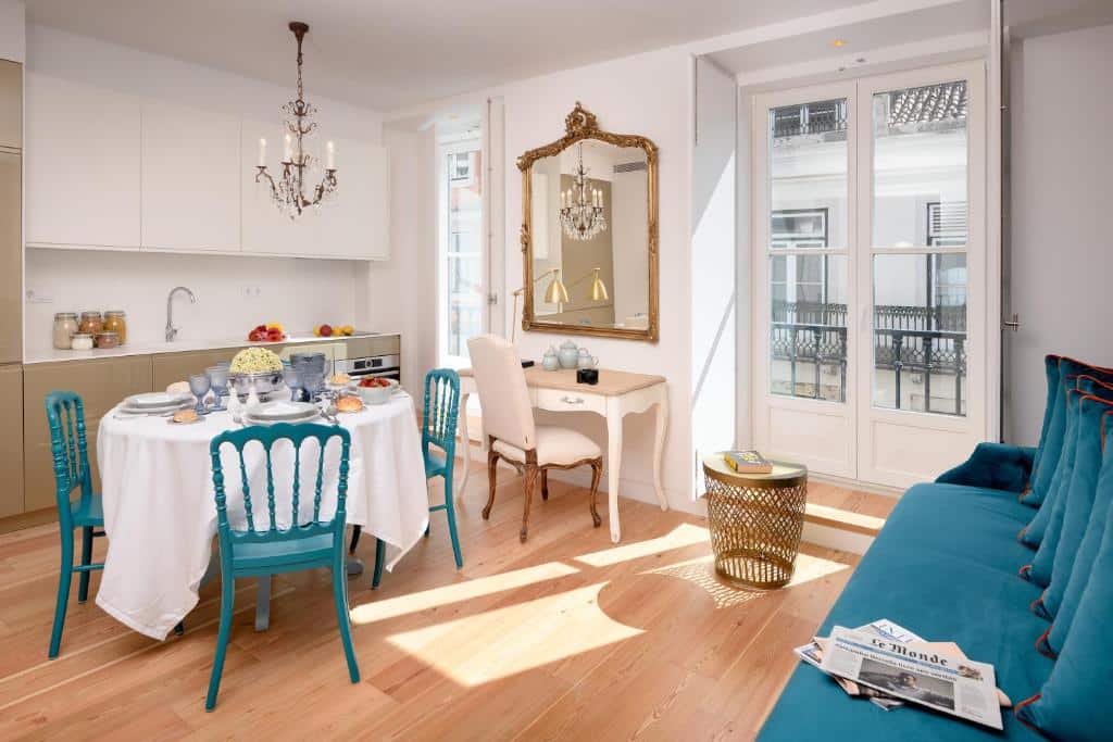 Sala de estar com cozinha integrada do Residentas Aurea, com uma pequena varanda e uma janela, um sofá com almofadas no canto direito e, do lado esquerdo, uma mesa redonda com quatro cadeiras e uma cozinha compacta com armário, placa e lava-loiça