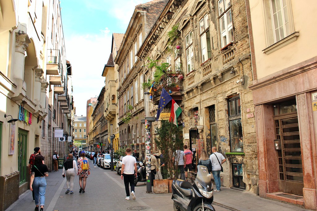 Pessoas a caminhar por uma rua estreita repleta de edifícios antigos, lojas e veículos estacionados, sob um céu parcialmente nublado, em Erzsébetváros.
