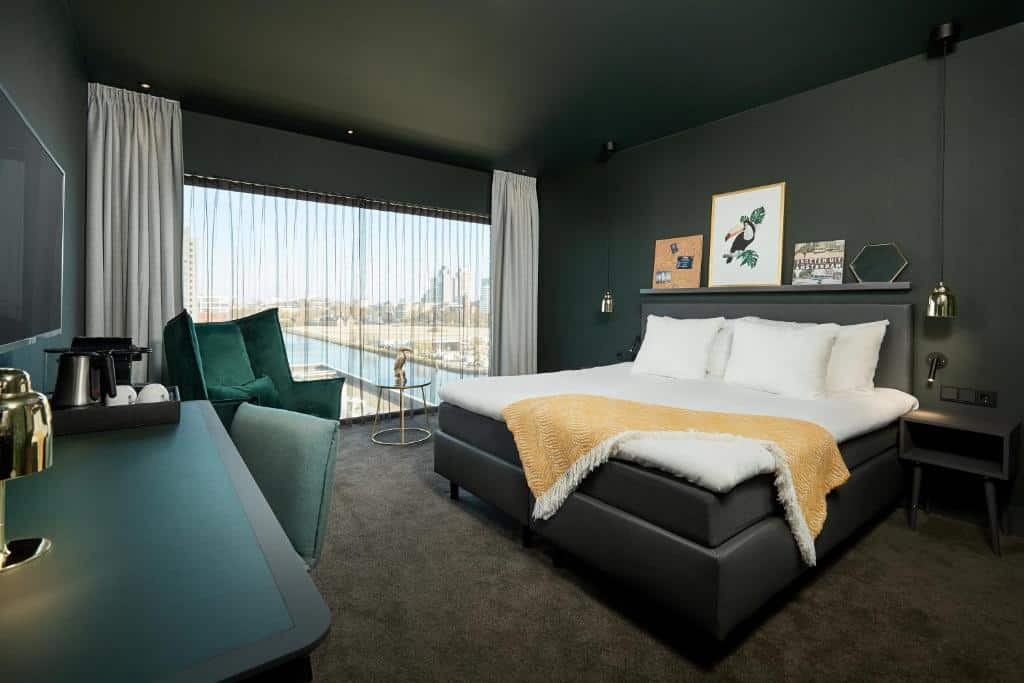 Quarto Comfort do Van der Valk Hotel, de 31 m², com cama de casal, cadeirão verde-escuro, mesa com cadeira e janela panorâmica com vista para a cidade