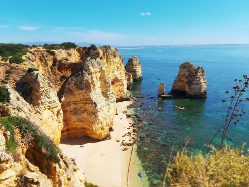 Vista da Ponta da Piedade, em Lagos, com formações rochosas douradas a erguerem-se do mar azul-cristalino e uma pequena praia.