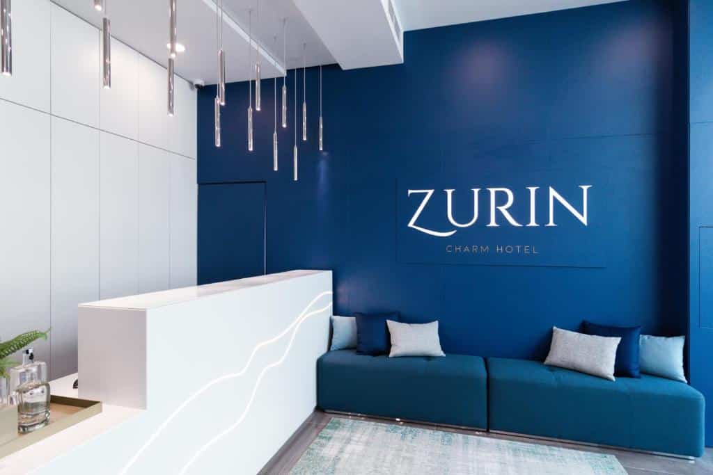 Receção de hotel moderna com parede azul, balcão branco e a placa Zurin Charm Hotel na parede.