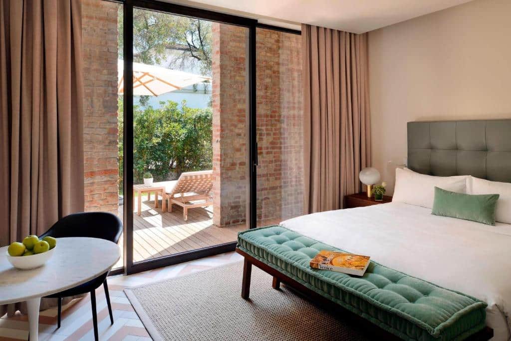 Quarto de hotel acolhedor com grandes janelas de vidro que se abrem para uma zona exterior com espreguiçadeiras e vegetação
