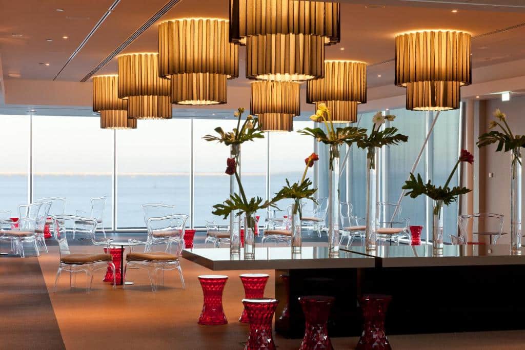 Área de refeições do MYRIAD by SANA Hotels com janelas panorâmicas viradas para o rio Tejo, lustres, cadeiras transparentes e arranjos de flores e vasos vermelhos