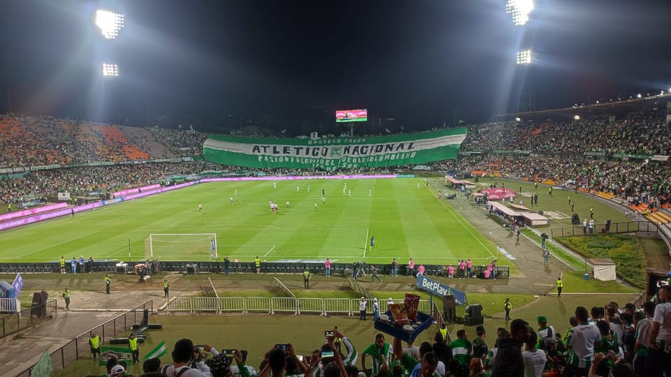 Jogo de futebol num estádio cheio de adeptos, com uma grande faixa do Atlético Nacional nas bancadas.