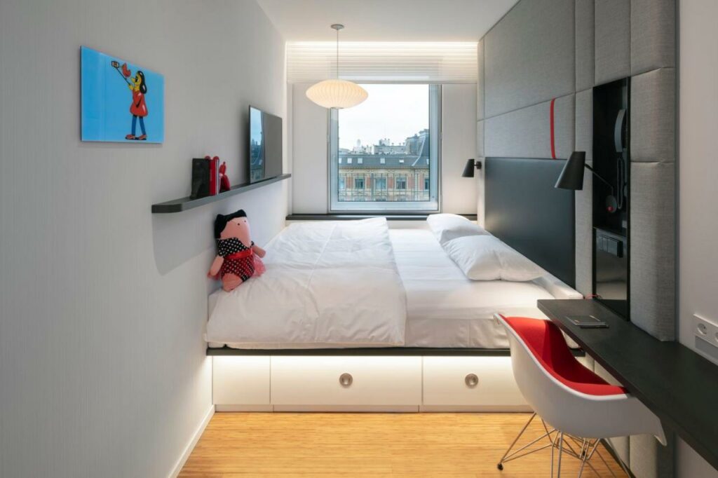 Quarto do citizenM Copenhagen Radhuspladsen com uma cama grande de casal com cabeceira acolchoada, uma prateleira com televisão e uma mesa estreita com cadeira. Tem também uma janela.