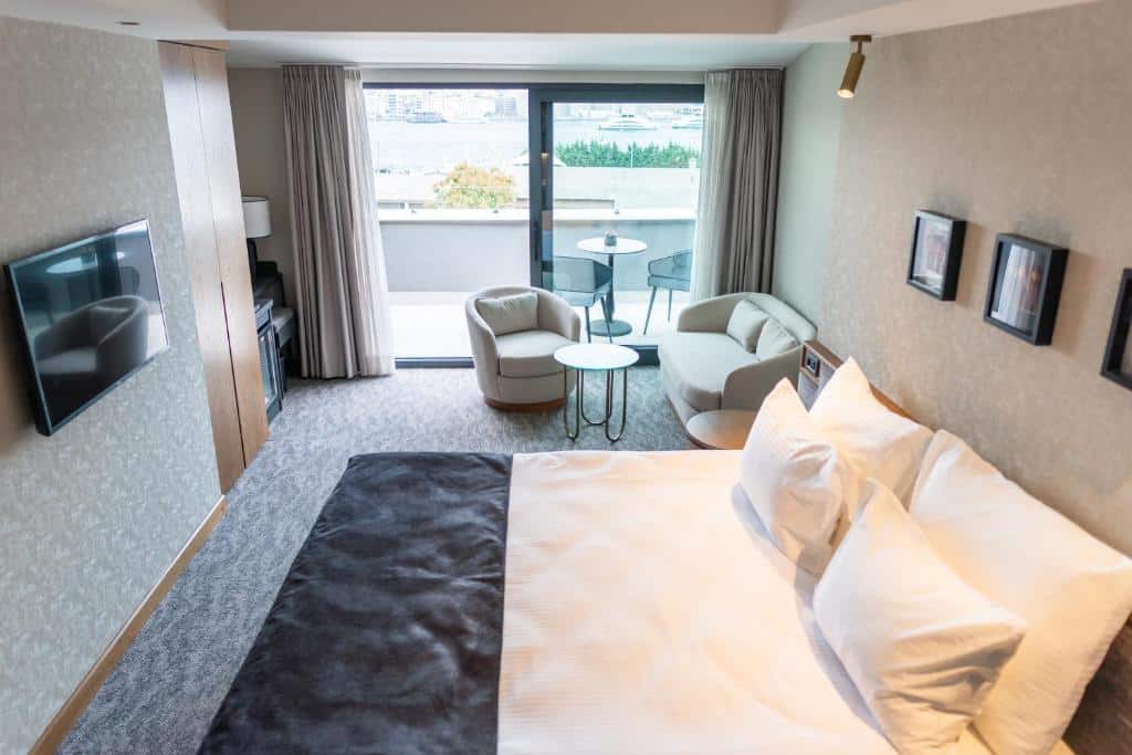 Quarto do Meg Hotel com uma cama grande, duas cadeiras, uma mesa, TV na parede e uma varanda com vista para o exterior.
