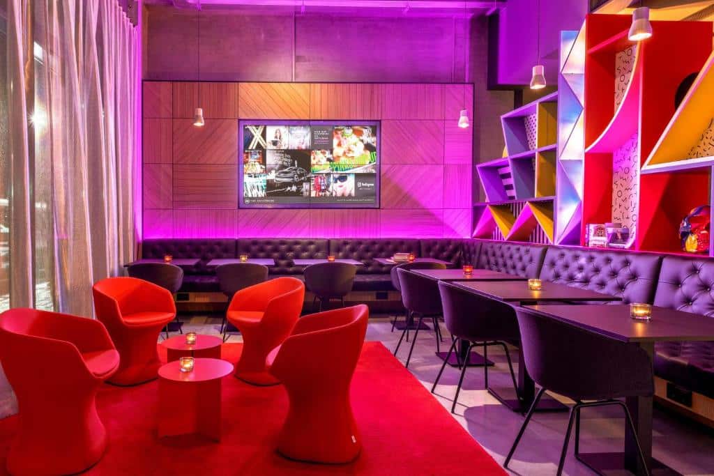 Lounge partilhado do Moxy Milan Linate Airport com poltronas vermelhas, sofás roxos, mesas quadradas e iluminação em tons de amarelo e rosa