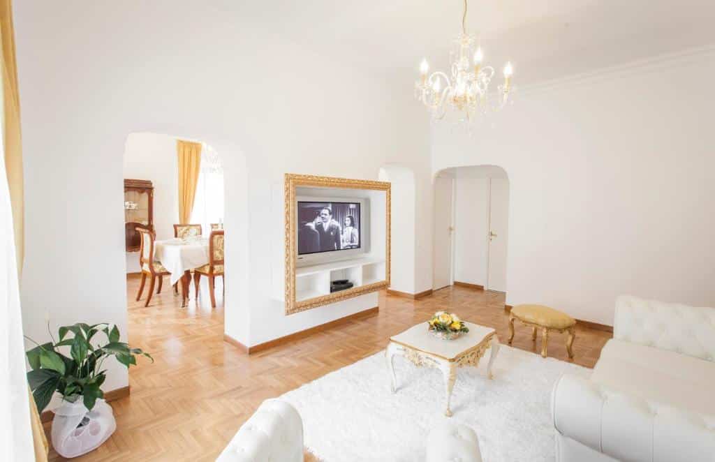 sala da Suite Sarandrea, com tapete, mesa de centro e sofás brancos, televisão em frente, num ambiente amplo em U ligado a uma cozinha com mesa e cadeiras e um lustre por cima