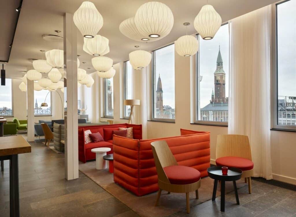 Zona comum do citizenM Copenhagen Radhuspladsen, com várias cadeiras, sofás e mesas. Há vários candeeiros diferentes a decorar o espaço e janelas com vista para a cidade.