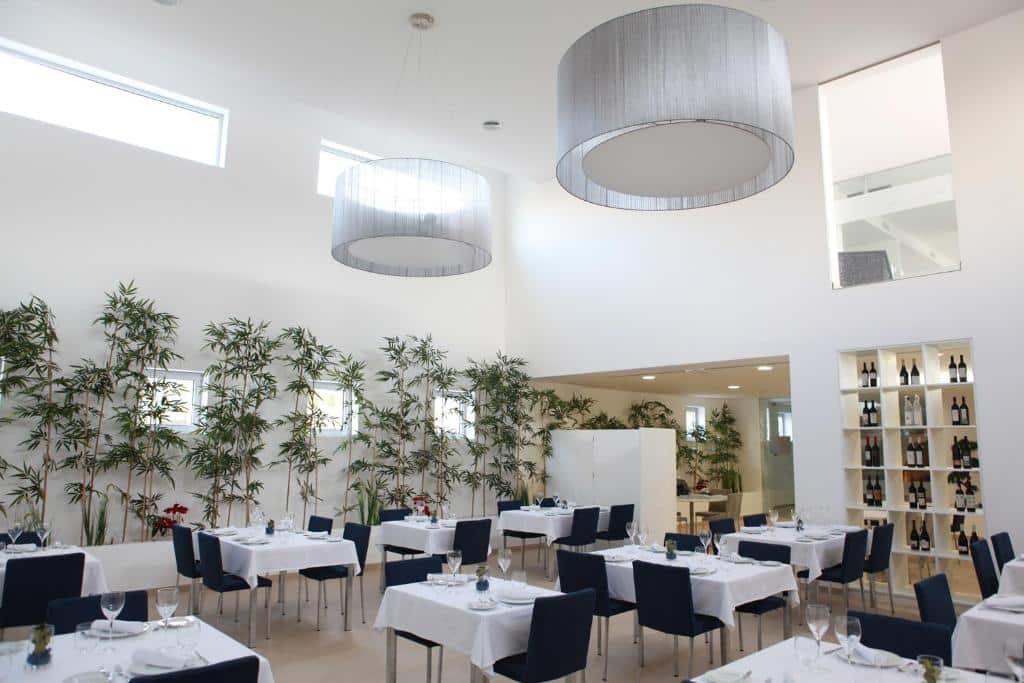 Sala de jantar de restaurante moderno com paredes brancas, mesas com toalhas brancas, cadeiras escuras e plantas numa parede, no Hotel Anjo de Portugal.