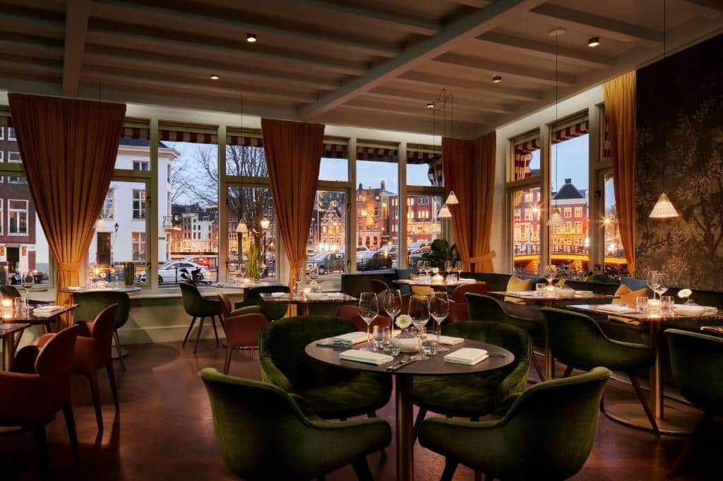 Sala de jantar de um restaurante elegante do Tivoli Doelen Amsterdam Hotel, com mesas redondas, cadeiras verdes postas para o jantar e janelas grandes com vista para uma rua da cidade.