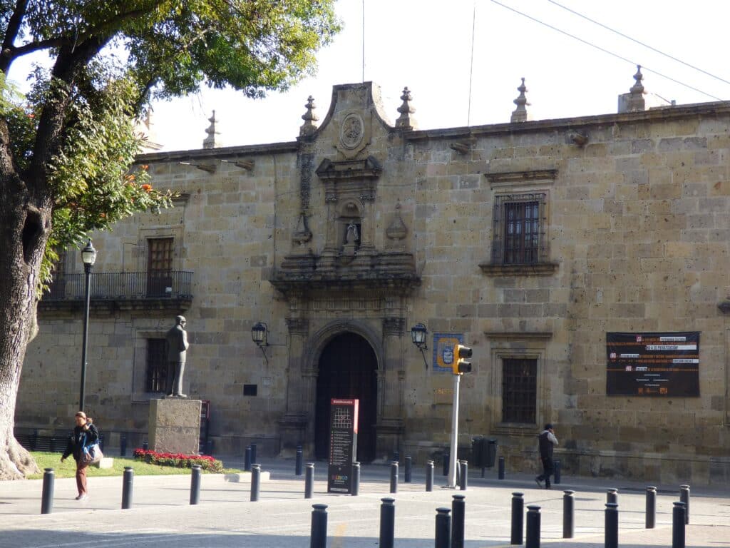Museu Regional de Guadalajara, edifício histórico em pedra com elementos arquitetónicos ornamentados, incluindo torres pontiagudas, numa praça com jardins e pessoas a circular.