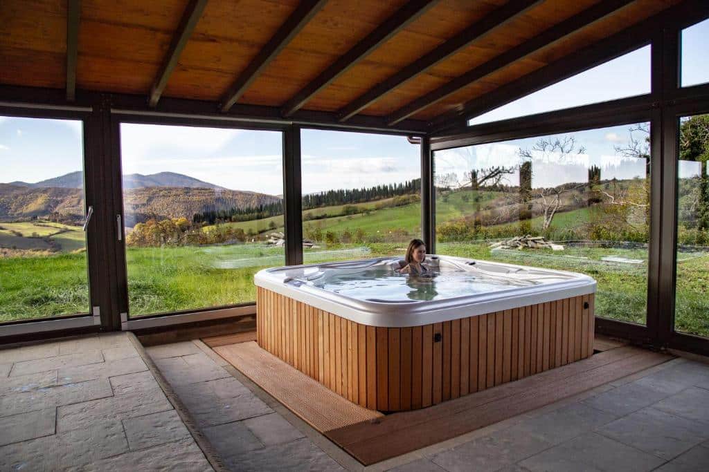 Espaço do Agri Resort &amp; SPA Le Colline del Paradiso, com paredes de vidro a mostrar uma paisagem verde e as montanhas da região, e uma mulher de cabelo curto dentro de uma hidromassagem quadrada