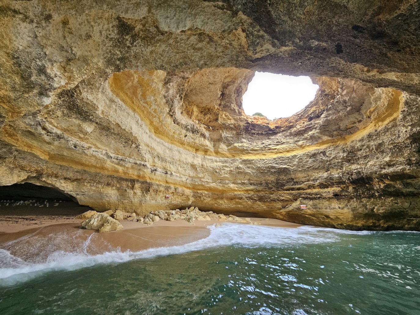 Gruta com uma grande abertura circular no topo, praia de areia no interior e ondas a chegar à costa, iluminadas por luz natural.