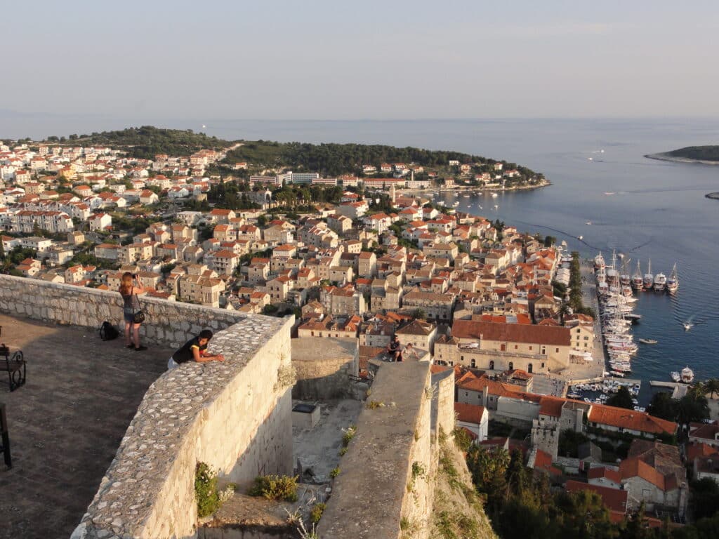 Vista aérea do Forte Espanhol de Hvar, com edifícios de telhados vermelhos, um porto com vários barcos e um muro de pedra em primeiro plano.