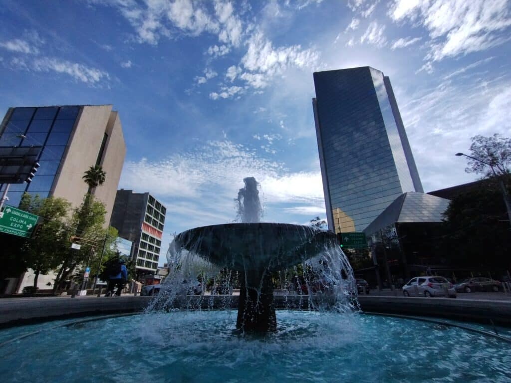 Fonte de água com repuxo central numa praça de cidade moderna, com edifícios altos ao fundo, na Avenida Chapultepec.