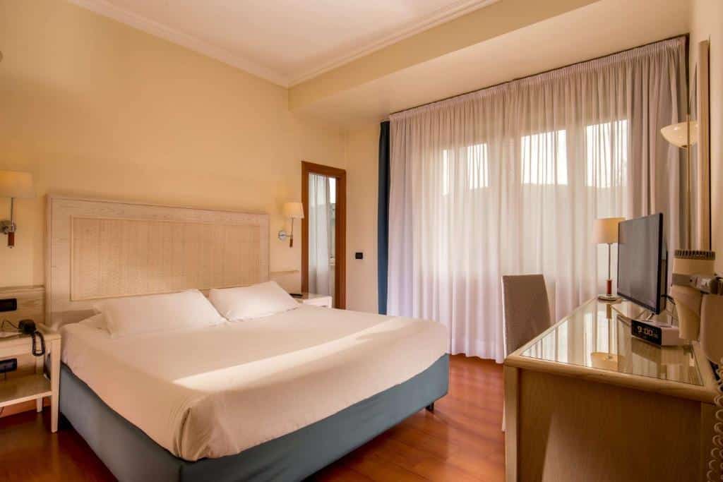 quarto do Best Western Hotel Globus, um dos hotéis baratos em Roma, com cama de casal grande, candeeiro e mesa de ambos os lados, uma mesa com cadeira espelhada e televisão em frente, e uma janela grande com cortinados leves e vista