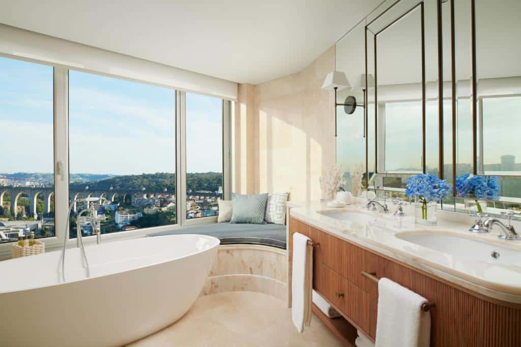 Casa de banho do Corinthia Lisbon com uma janela enorme com vista para a cidade, um pequeno sofá junto à janela, uma banheira oval e um lavatório com duas cubas e um amplo espelho