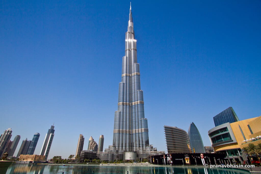 O Burj Khalifa, um arranha-céus alto e moderno contra um céu azul claro, rodeado por edifícios mais baixos, com reflexo na água.