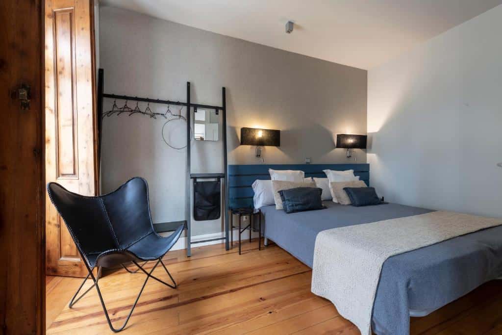 Quarto minimalista do Living Lisboa Baixa Apartments com cama de casal, roupa de cama cinzenta, chão de madeira, cadeira preta, apliques de parede e um pequeno bengaleiro.