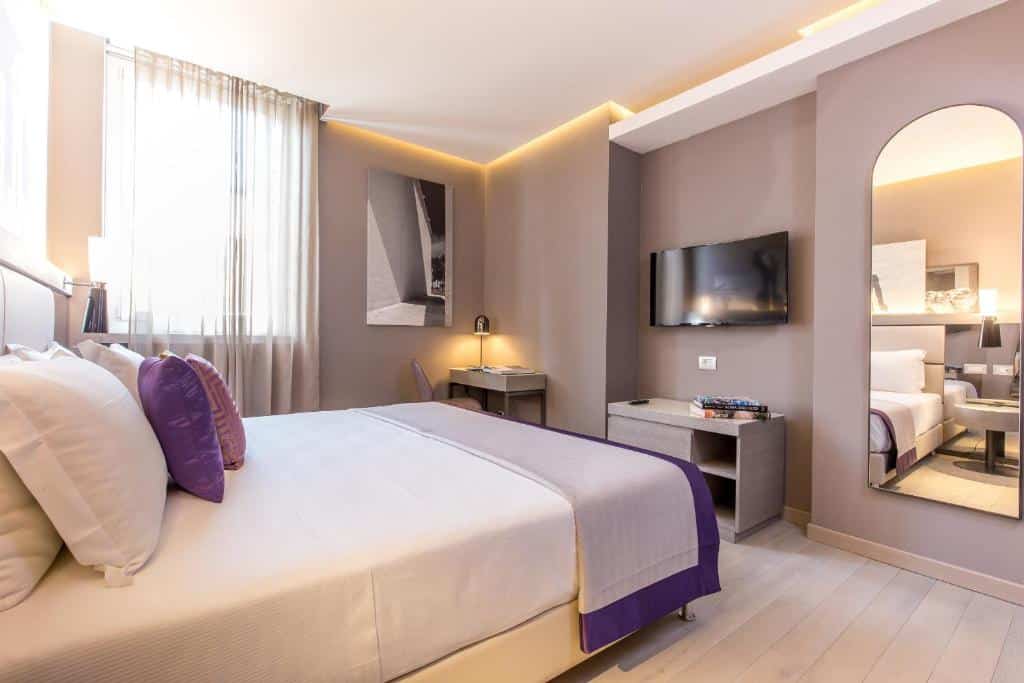 quarto do Orazio Palace Hotel, um dos hotéis perto do Vaticano, com cama de casal, cómoda e televisão em frente, mesa e cadeiras, janela com cortinas leves de um lado e um grande espelho do outro, em tons bege com pormenores roxos