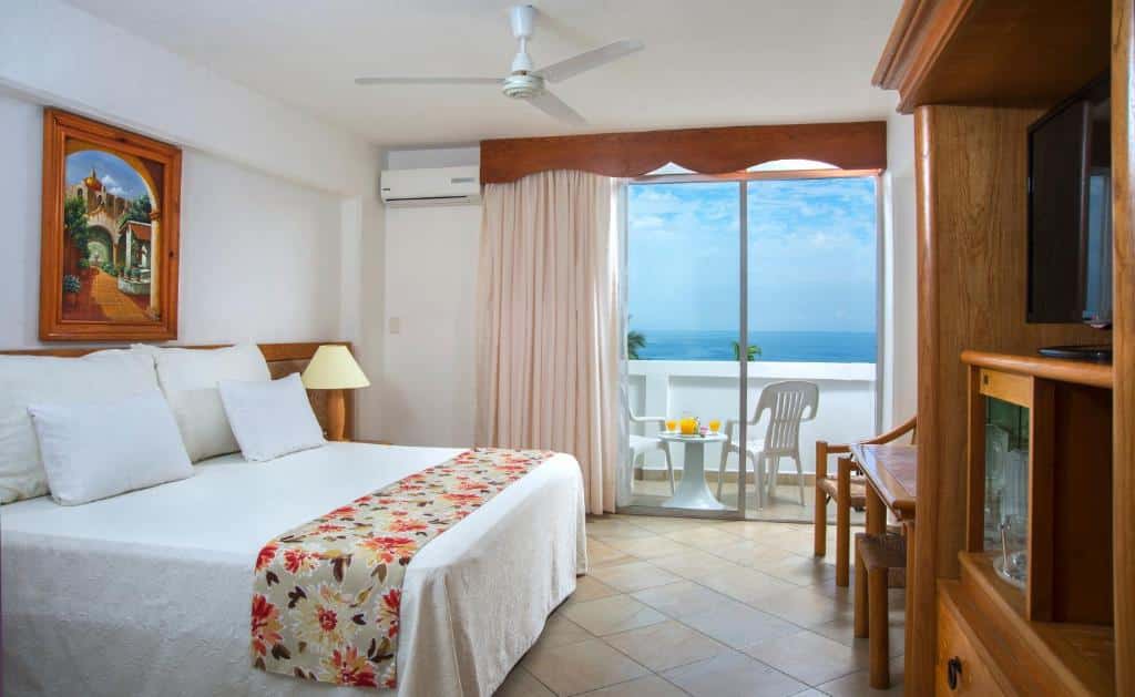 Quarto do Tropicana Hotel Puerto Vallarta com uma cama, colcha floral, mesa, cadeiras, TV e varanda com vista para o mar.