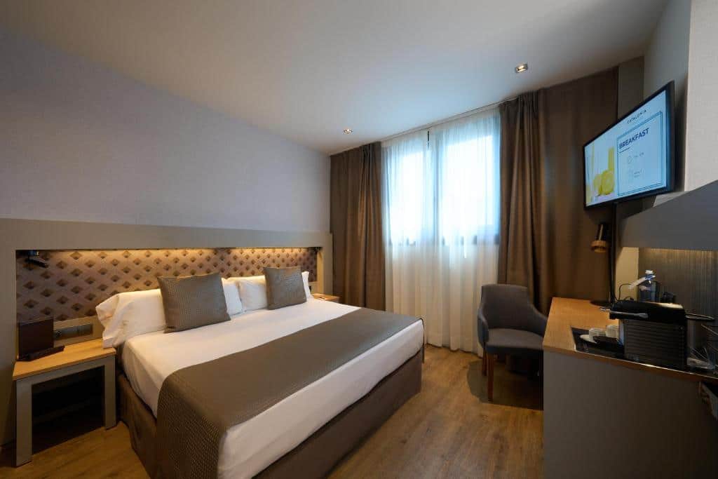 Quarto do Catalonia Port, uma das recomendações de hotéis no Bairro Gótico em Barcelona. A cama de casal encosta a uma cabeceira acolchoada, com mesas de cabeceira de ambos os lados e uma secretária com televisão em frente.