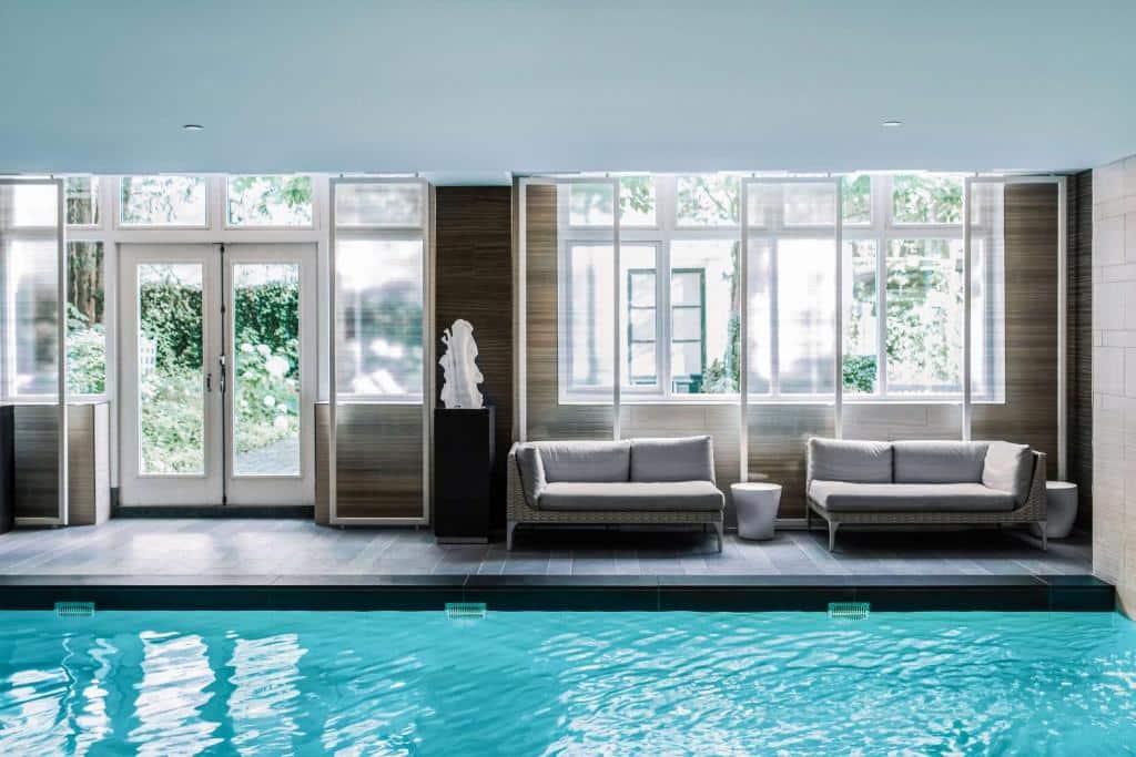Piscina interior do Waldorf Astoria Amsterdam, com água azul-clara, dois sofás cinzentos, mesas de apoio brancas e janelas grandes que deixam entrar luz natural.