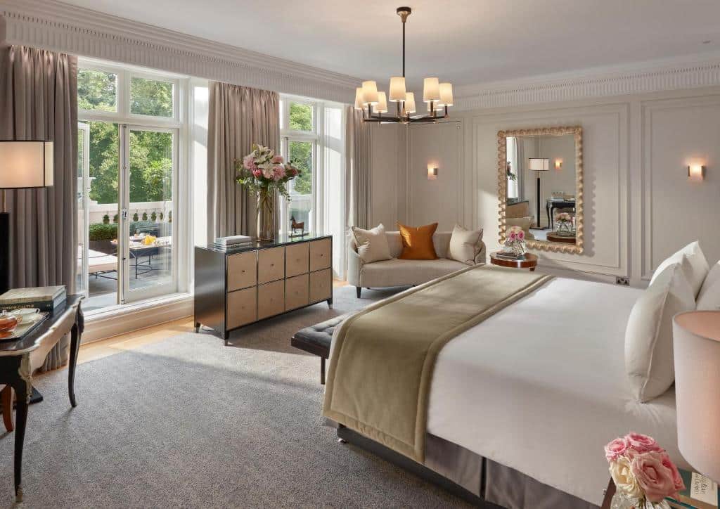 Quarto espaçoso do Mandarin Oriental Hyde Park com uma varanda ampla com vista para árvores e poltronas ao ar livre; no interior há uma cómoda, cama de casal, um sofá com almofadas, um lustre e um espelho.