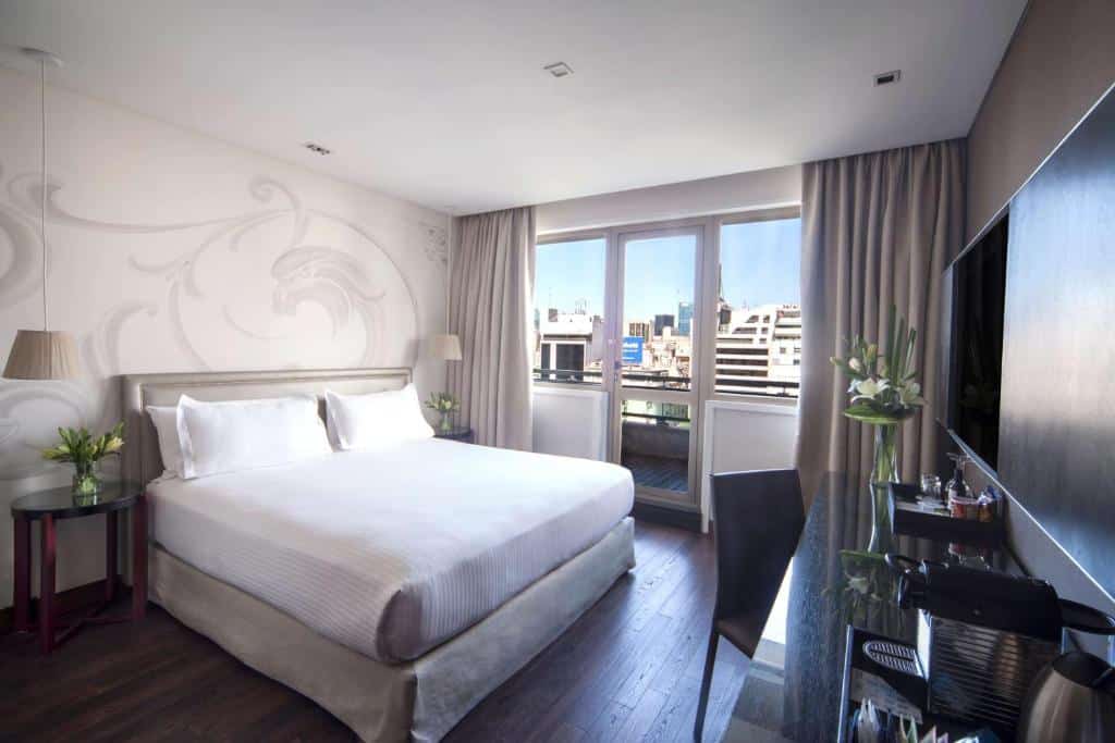 quarto do NH Buenos Aires Tango com cama de casal ao centro, mesas e candeeiros dos dois lados, secretária com cadeira e tv, e uma varanda com vista para a cidade, em tons claros com madeira escura