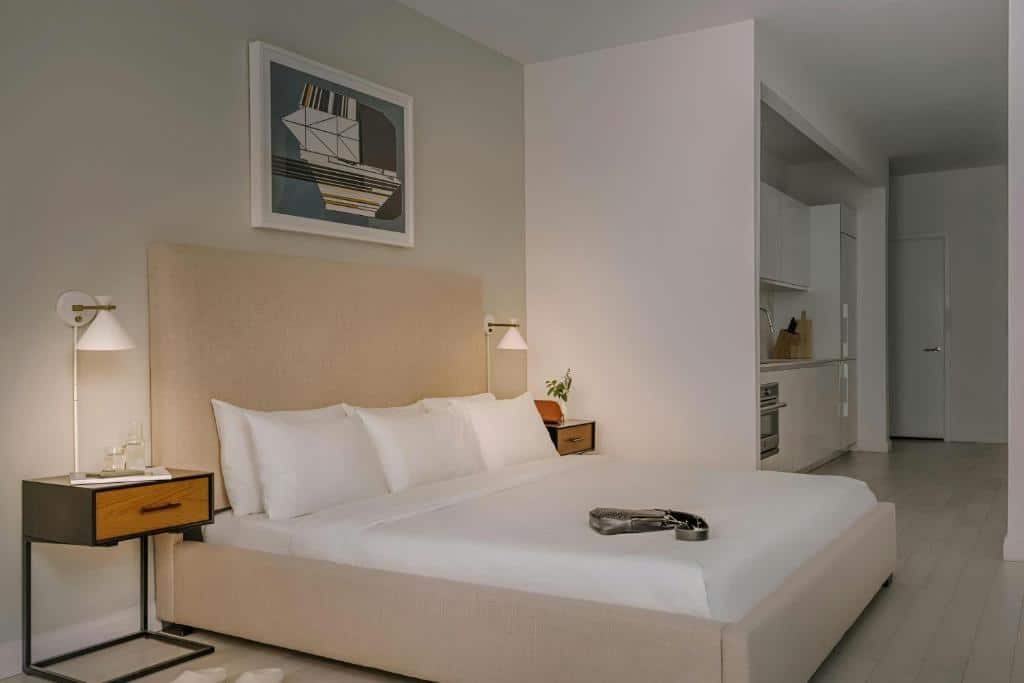Quarto moderno com uma cama bege, duas mesas de cabeceira, candeeiros de parede, arte abstrata e uma pequena zona de cozinha ao fundo.