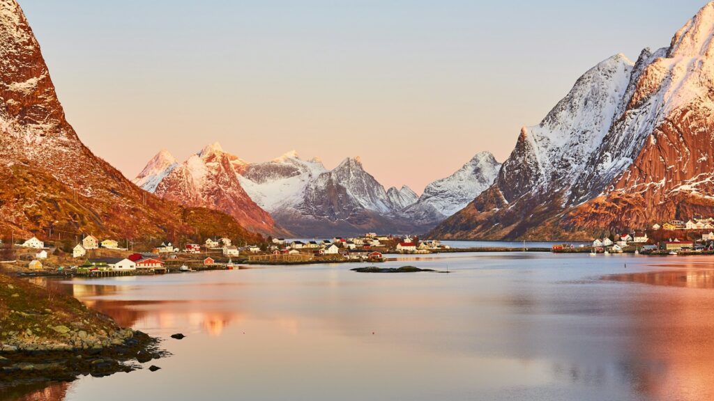 Paisagem de uma aldeia nas Ilhas Lofoten, na Noruega, durante o nascer do sol. As casas alinham-se ao longo da costa, refletindo-se nas águas. Montanhas cobertas de neve rodeiam a aldeia, banhadas pela luz dourada do sol.