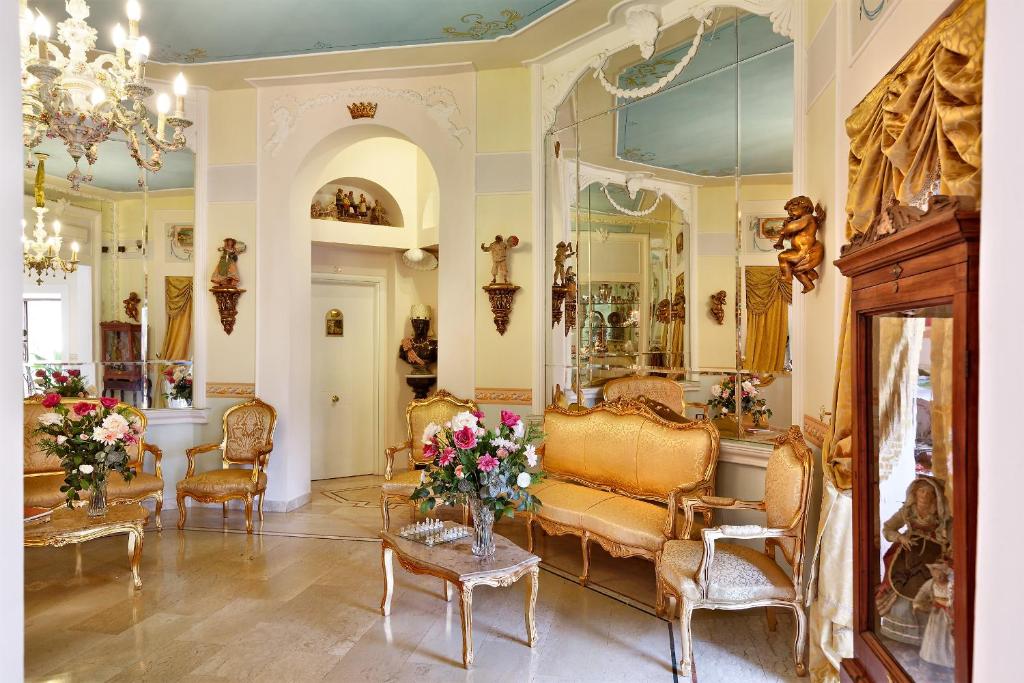 Sala de estar luxuosa do Hotel Art Resort Galleria Umberto, em estilo barroco, com pormenores dourados, espelho ornamentado, lustres de cristal e sofá e cadeiras estofados em tons dourados e florais.