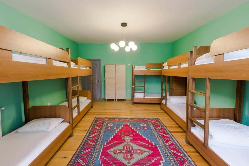 Um quarto com seis beliches de madeira, um tapete colorido, paredes verdes, um roupeiro branco e um armário ao fundo.