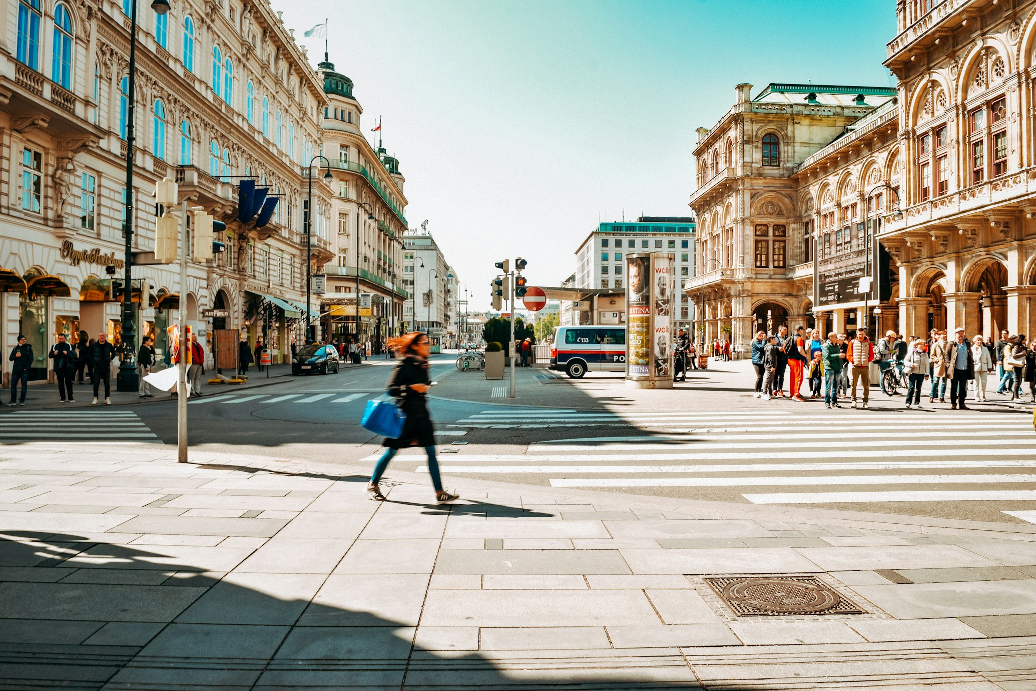 Cruzamento movimentado no centro de Viena, com pessoas a atravessar a rua e edifícios ornamentados em redor.