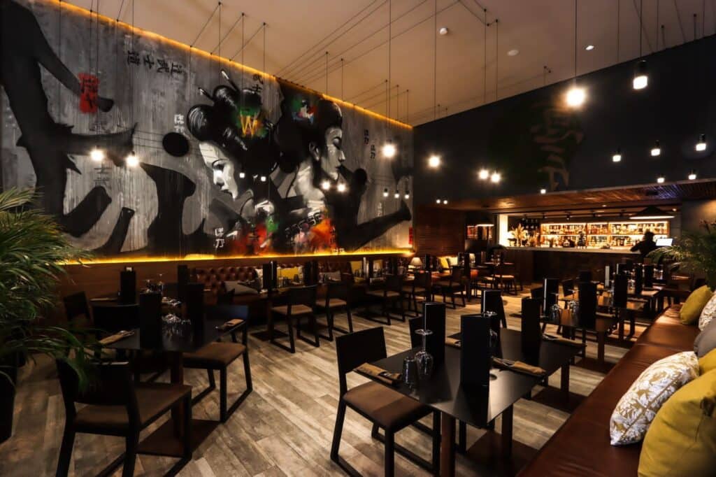 O restaurante Poka com pouca iluminação e decoração moderna, com um mural de gueixas na parede e mesas com ementas e copos, em Marraquexe.