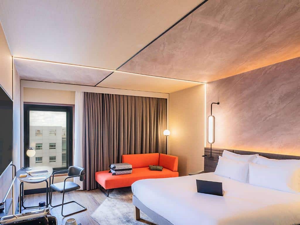 Quarto do Novotel Lisboa com cama branca, sofá vermelho, mesa redonda, cadeiras e janela grande com cortinas.
