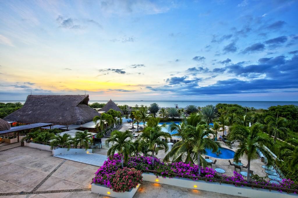 Vista panorâmica de um resort tropical ao pôr do sol, com telhados de colmo, palmeiras, uma piscina e o mar ao fundo.