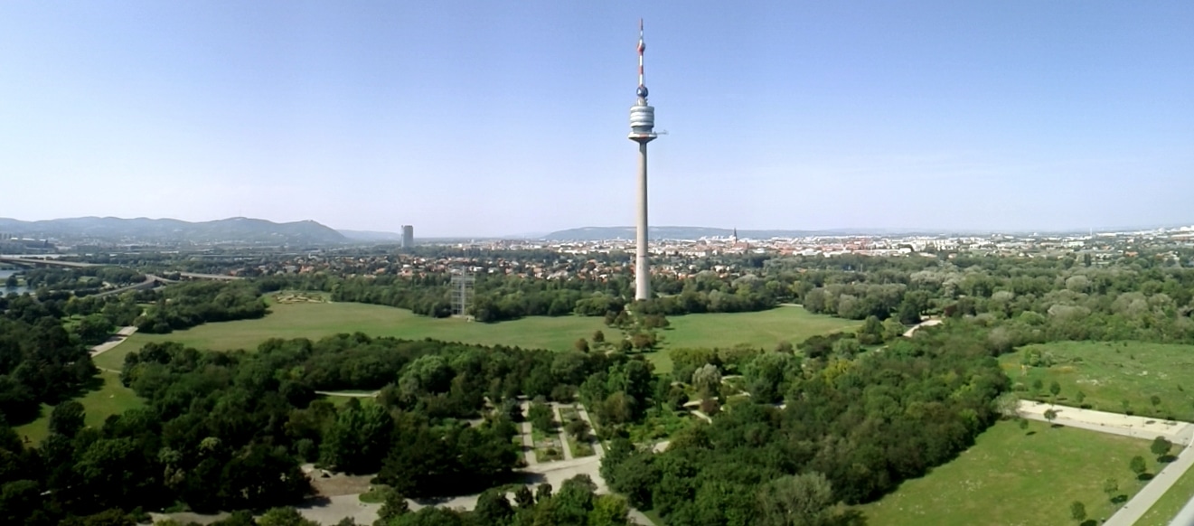 Vista panorâmica do Donaupark, com a alta torre do Danúbio a erguer-se entre relvados, caminhos e árvores, e a cidade ao fundo.