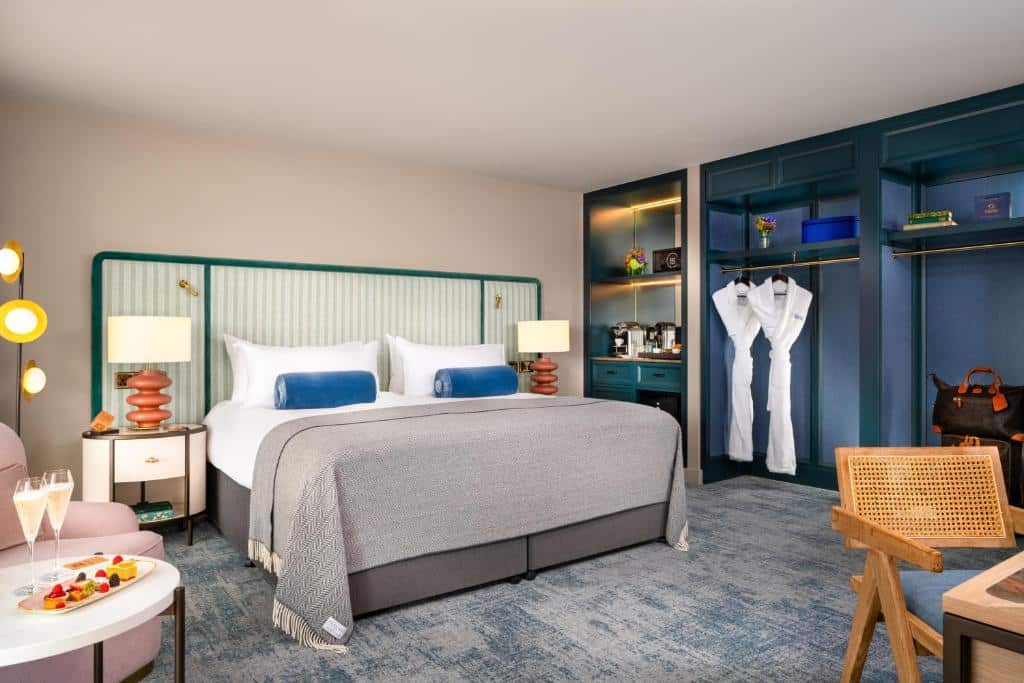 Quarto do The Green com decoração em tons de azul e verde, com cama de casal com roupa clara, mesas de cabeceira, roupeiro com prateleiras, poltrona de madeira e uma mesa com fruta e bebidas.