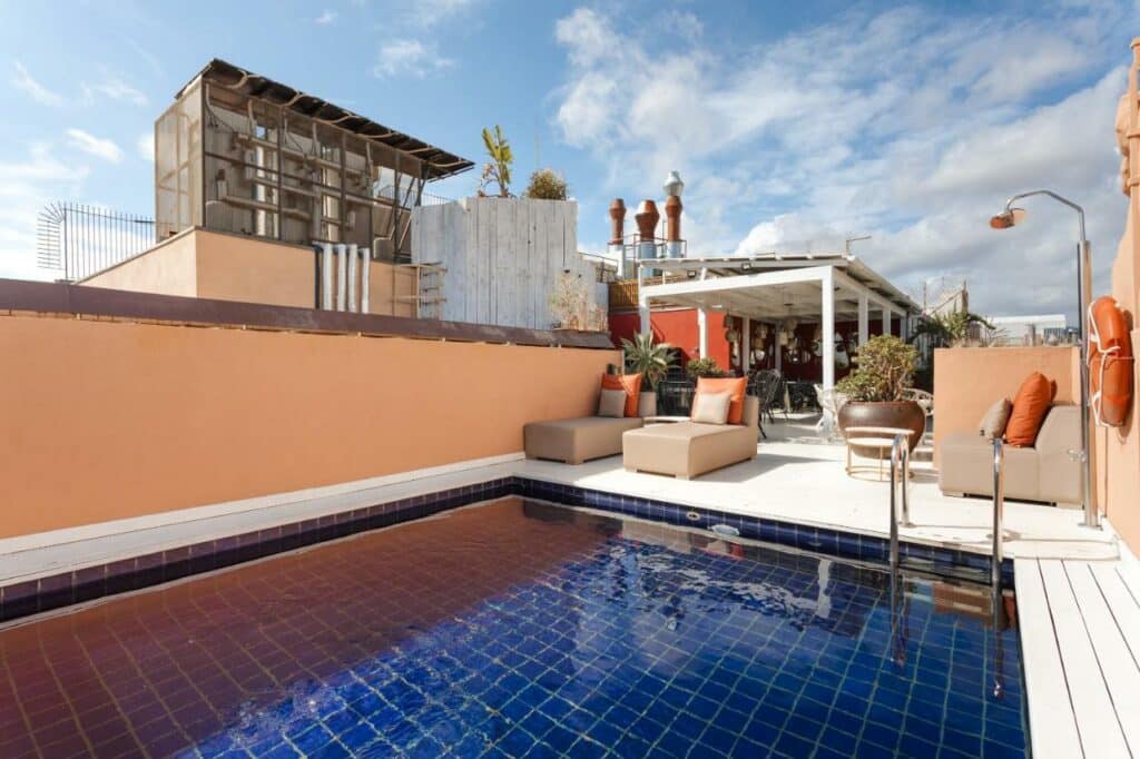 Zona da piscina do Sonder DO Plaça Reial, com espreguiçadeiras acolchoadas, chuveiro e uma área coberta com plantas ao fundo.