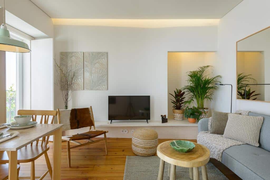 Sala de estar de um dos apartamentos do Dona Graca Lisbon Apartments com uma televisão, alguns vasos de plantas, um espelho amplo na parede do lado direito onde há também um sofá e uma mesa de centro de madeira rústica, além de uma mesa com dois lugares e uma janela ampla