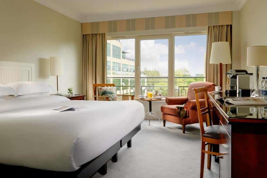 Quarto do Herbert Park Hotel and Park Residence com janelas amplas e vista para edifícios ao fundo. A cama tem roupa branca, e há um sofá castanho-claro e uma secretária com cadeira de madeira ao lado.