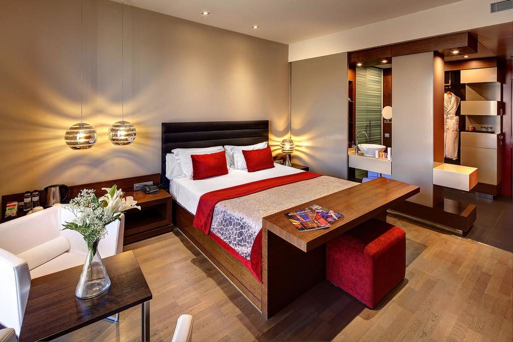 Quarto do Olivia Plaza Hotel, uma das recomendações de hotéis no Bairro Gótico em Barcelona. A cama tem lençóis e almofadas brancos e vermelhos, com uma mesa de madeira aos pés e candeeiros nas mesas de cabeceira.