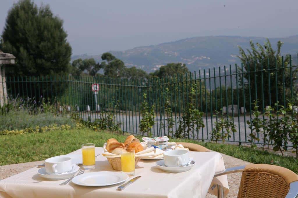 Mesa de pequeno-almoço ao ar livre no Hotel do Lago, com sumo, bolos e chávenas, e vista para as colinas verdejantes.