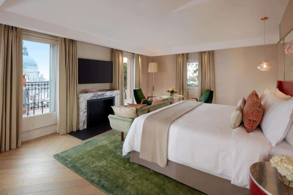 Quarto com janelas e varanda num hotel de luxo, com cama de casal, poltronas verdes, lareira e decoração elegante em tons neutros