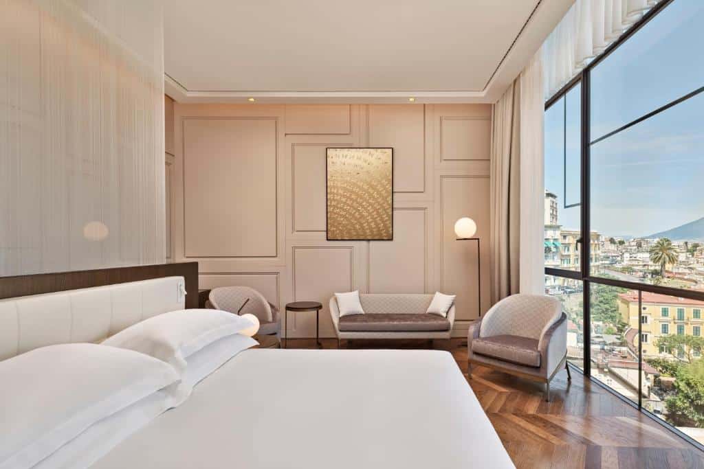 Quarto elegante do De Bonart Naples com mobiliário bege, quadro dourado na parede e vista para uma cidade litoral com edifícios e vegetação.
