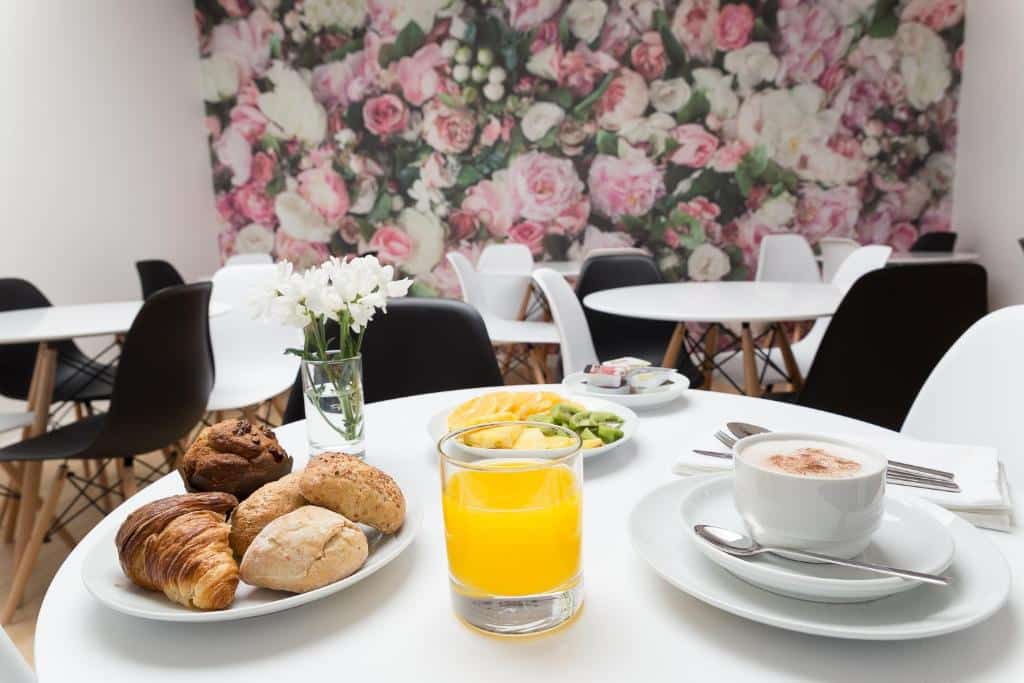 Uma mesa com bolos, sumo de laranja, fruta e café numa sala com papel de parede floral e cadeiras brancas, servida no Hotel Santa Isabel.